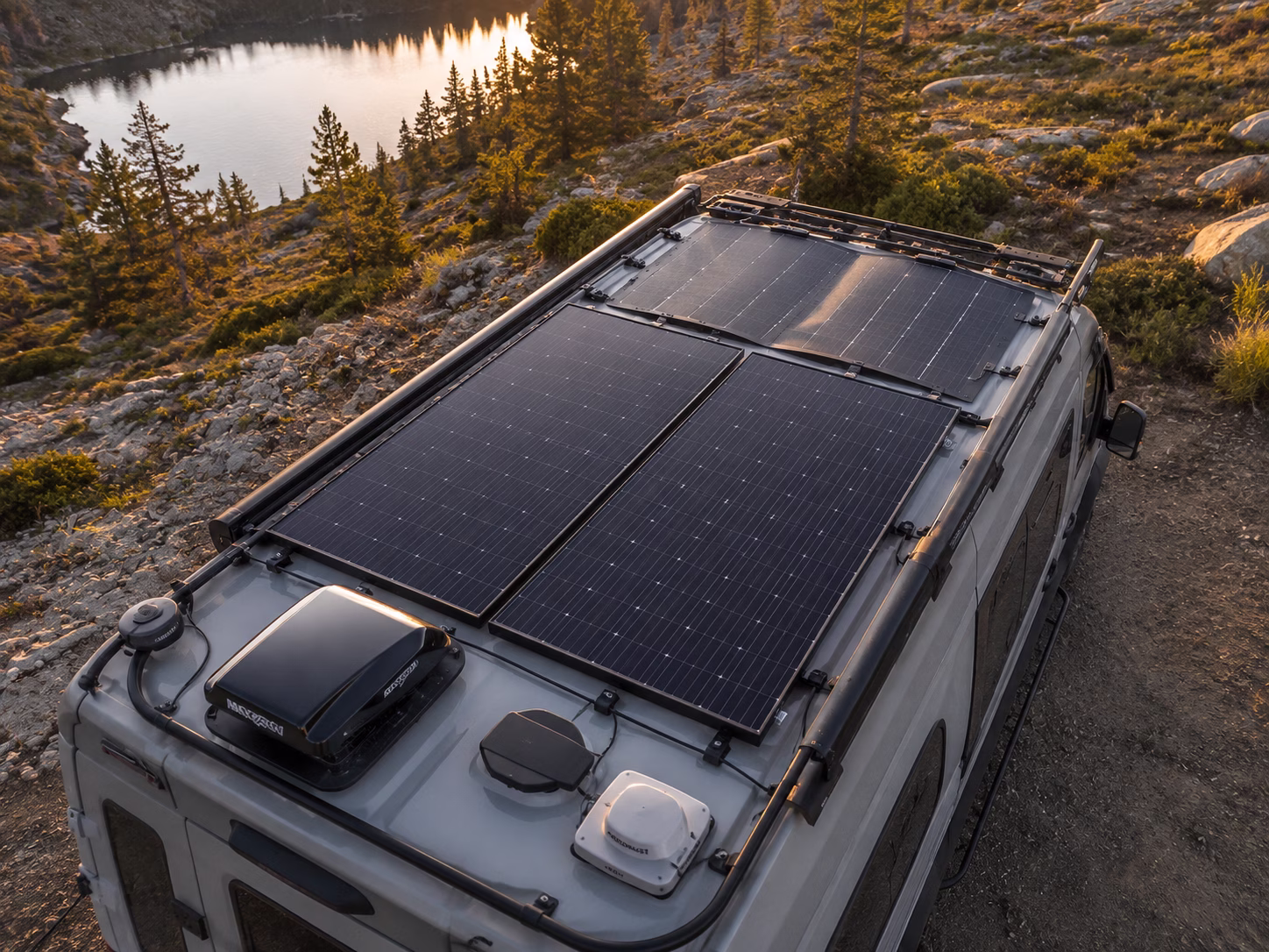 Aerial view of a rugged camper van with a large, modern solar panel array on the roof, parked in a scenic mountain landscape during golden hour.